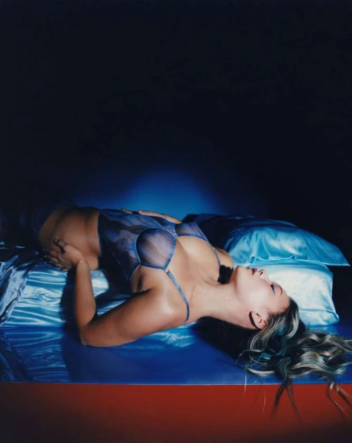Adult female model poses on a bed with blue satin sheets. She is wearing a blue lingerie set in a dimly lit indoor environment.