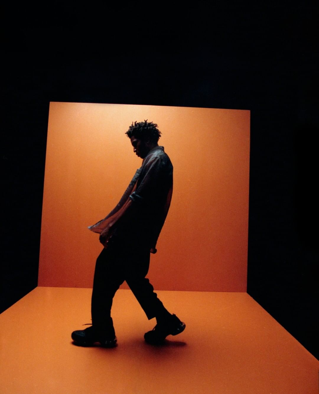 Silhouette of a stylish adult male walks across an orange stage in a dark studio with black shoes and a blue denim shirt.