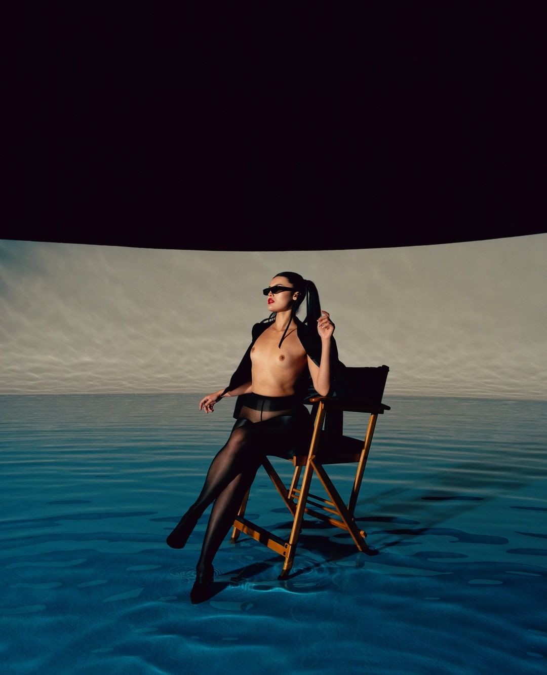 Adult woman with sunglasses poses topless on a director's chair in front of a water and sky backdrop, studio setting, striking light.