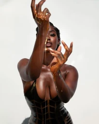 Black woman wearing a glossy dark bodysuit posing with honey dripping from her raised hands against white