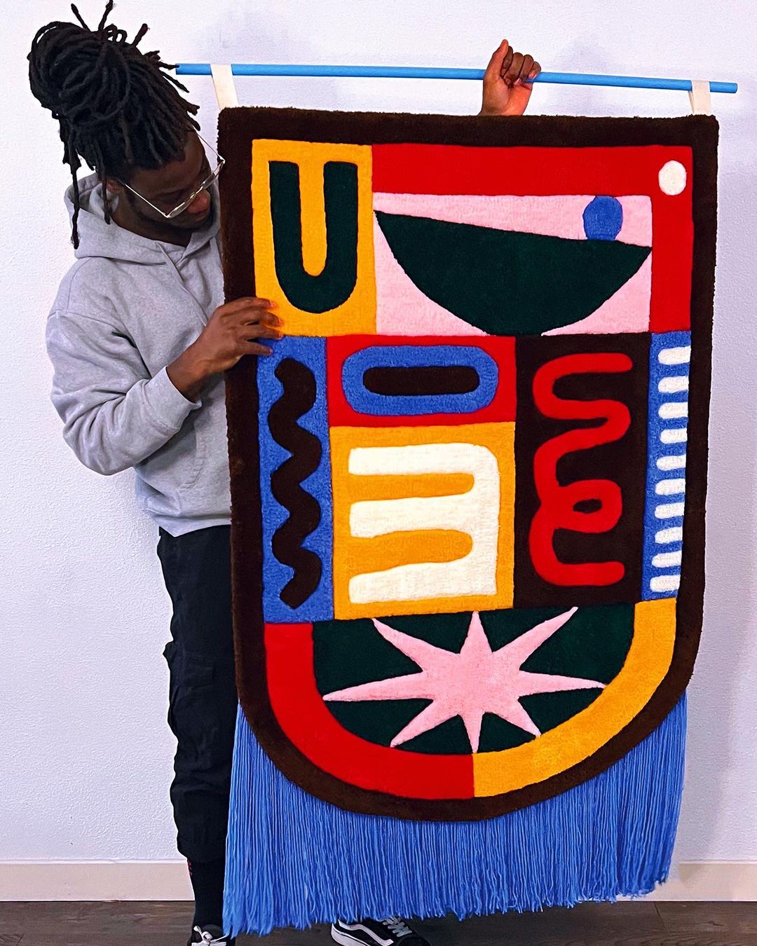 A person displays a colorful, geometric fiber art tapestry hanging from a blue rod. The tapestry features shapes and fringe.
