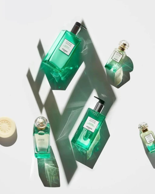 Five green bottles of Hermès Eau de Basilic perfume, soap and hand gel are arranged on a white surface with geometric shadows.