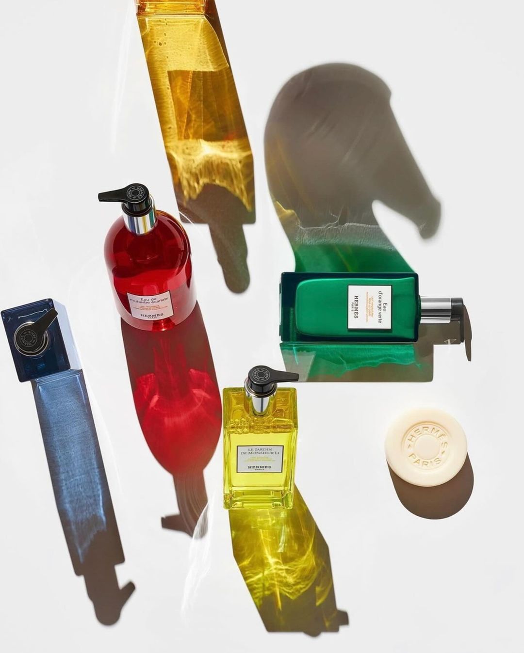 Arrangement of colorful Hermès bath and body products, including soap, hand and body cleansing gel, and shadows of the products on white.