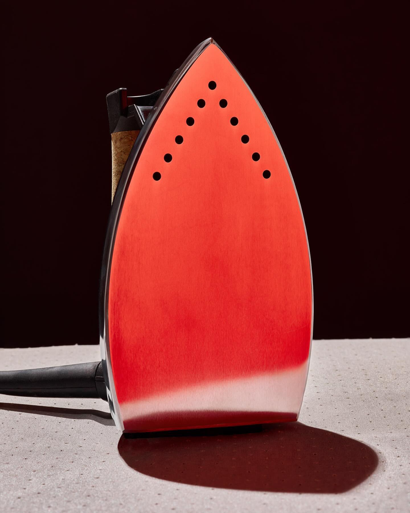 Close-up studio shot of a modern red and silver electric iron sitting on a grey surface against a dark, blurred background.