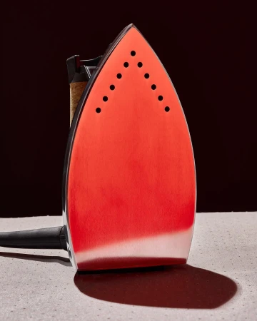 Close-up studio shot of a modern red and silver electric iron sitting on a grey surface against a dark, blurred background.