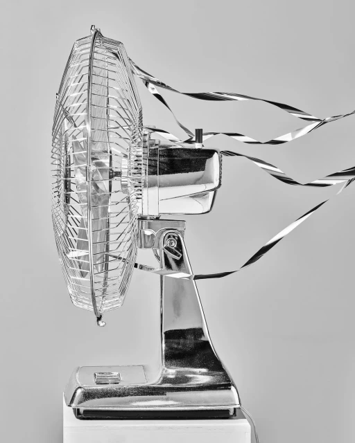 Black and white photo of a retro chrome electric fan on a white surface with reflective ribbon blowing around it, against a grey background.