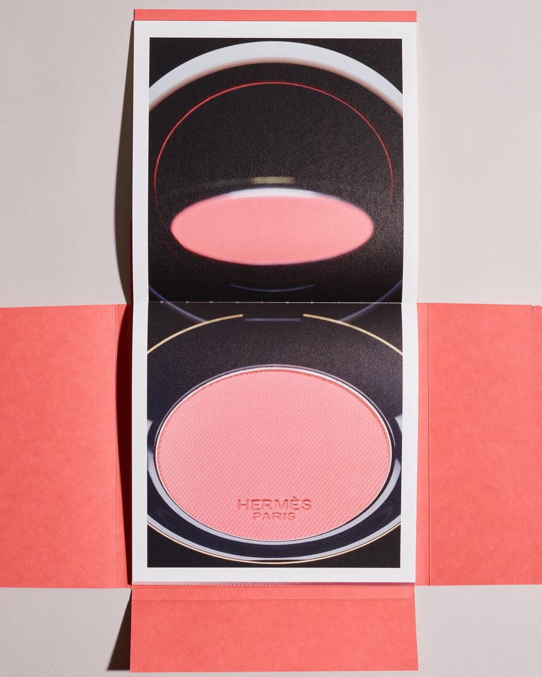 Hermes Paris blush compact. The cosmetic is pink and nestled in a coral-colored paper folding design against a neutral backdrop.