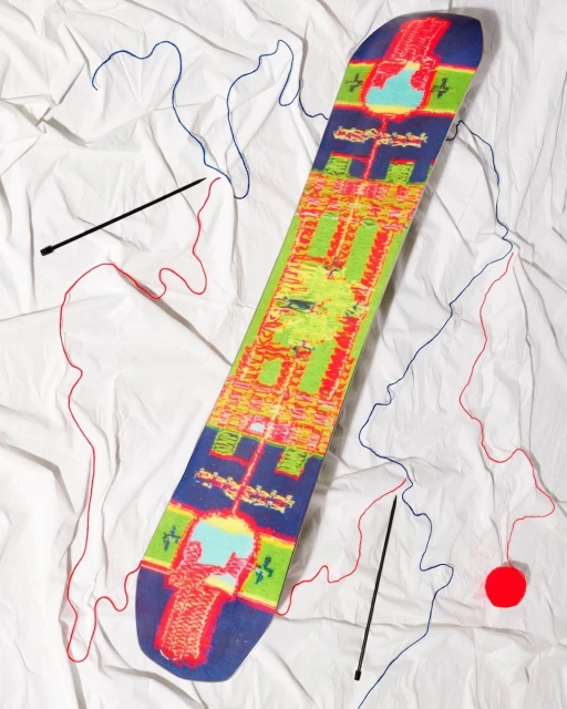 Overhead shot of a snowboard with a colorful pattern on a white crumpled sheet, with yarn and knitting needles scattered around it.