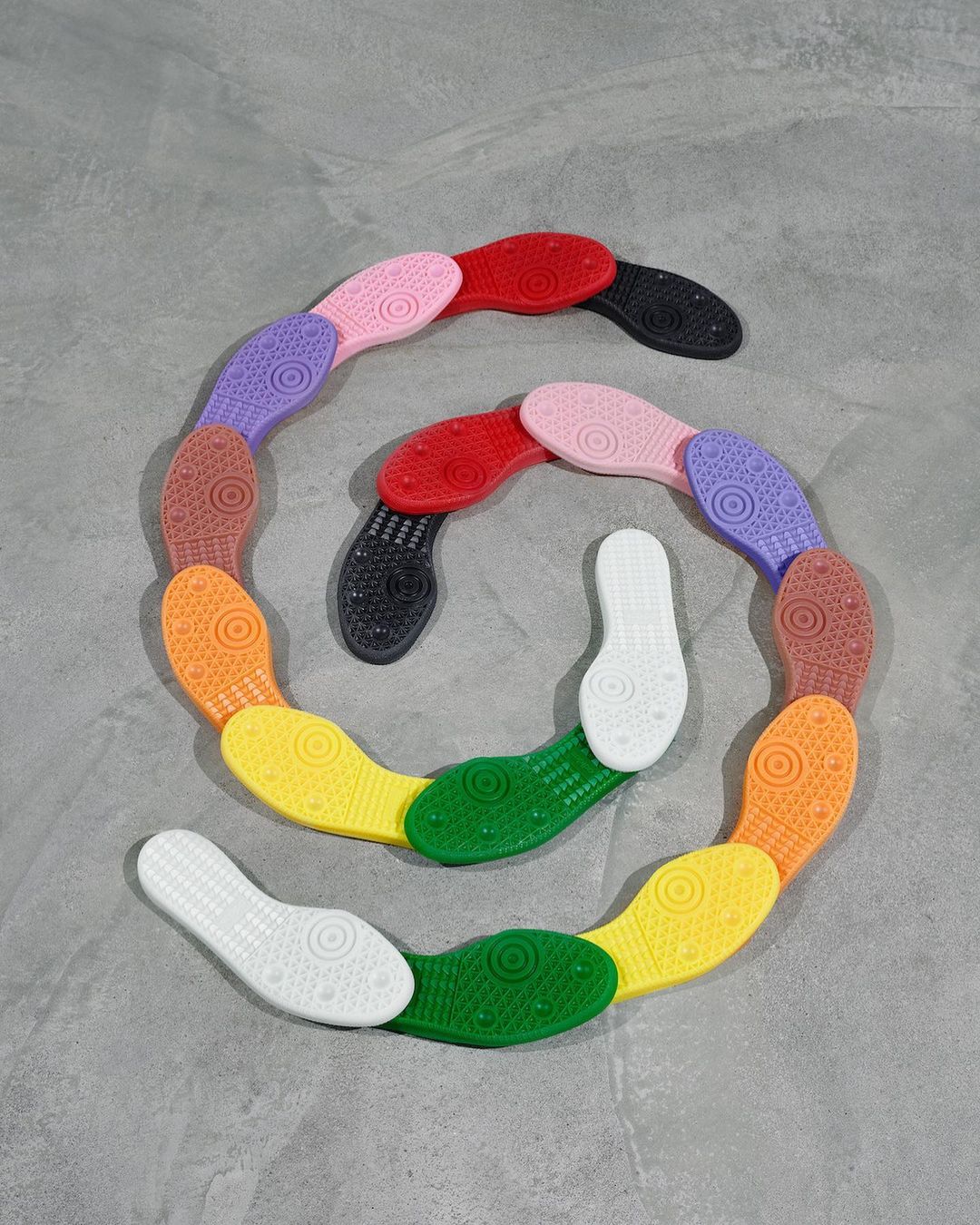 Colorful soles of shoes arranged in a spiral on a gray background, showcasing a spectrum of colors from white to black.