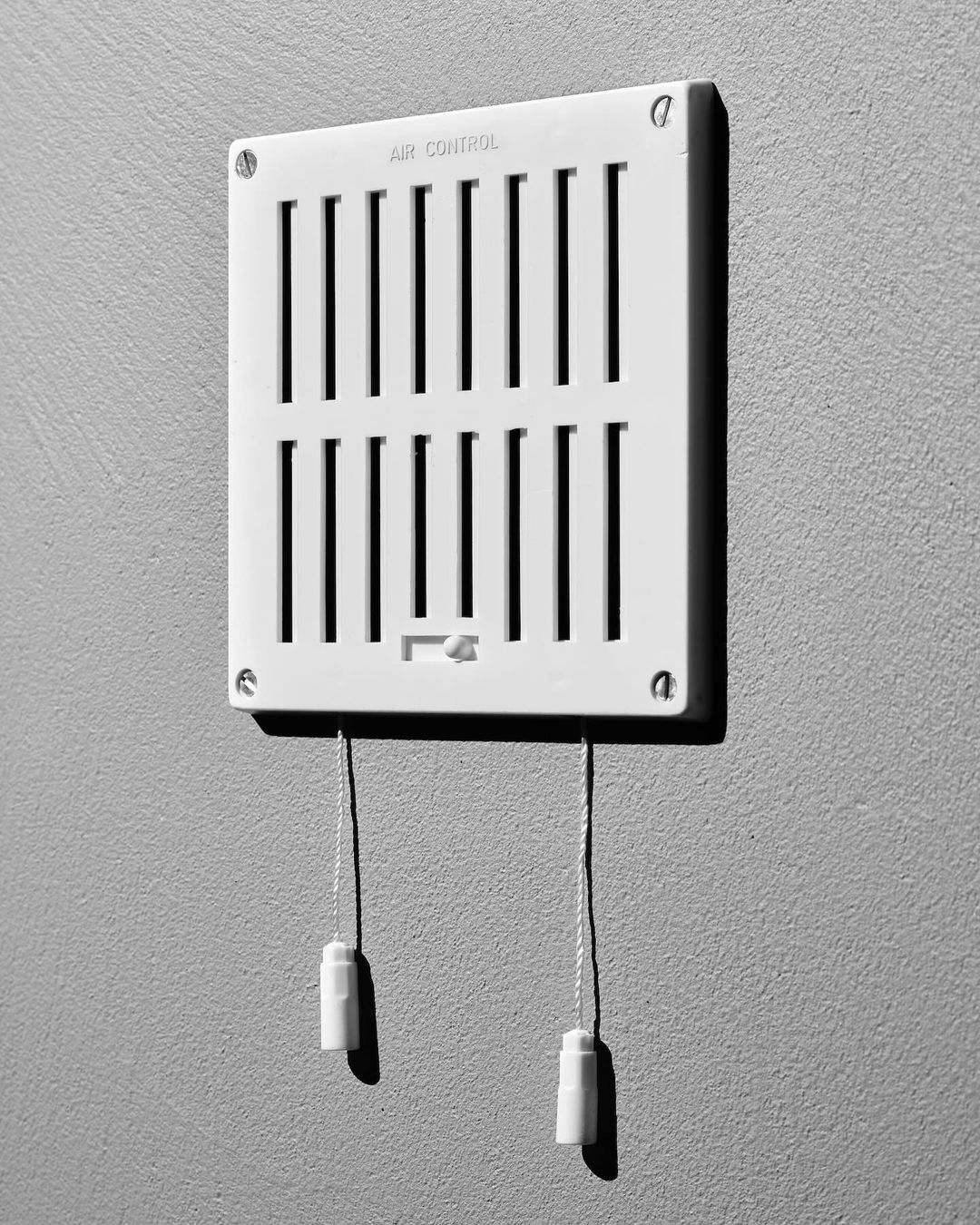 Black and white image of a white air vent on a gray wall. The vent has adjustable louvers and pull cords.