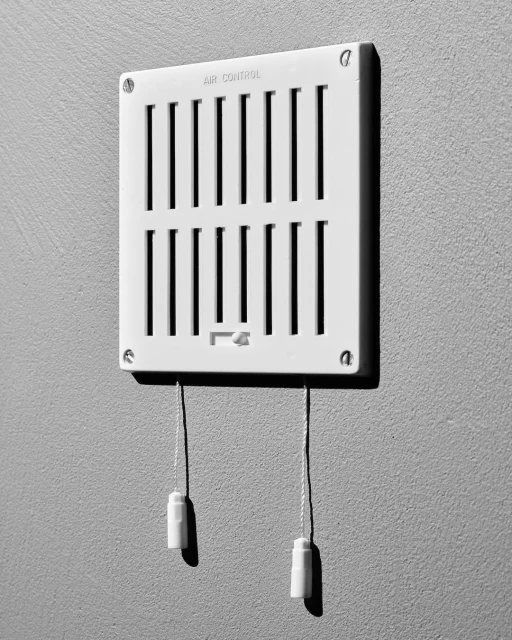 Black and white image of a white air vent on a gray wall. The vent has adjustable louvers and pull cords.