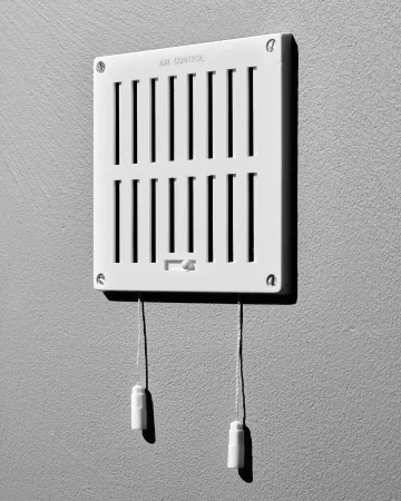 Black and white image of a white air vent on a gray wall. The vent has adjustable louvers and pull cords.
