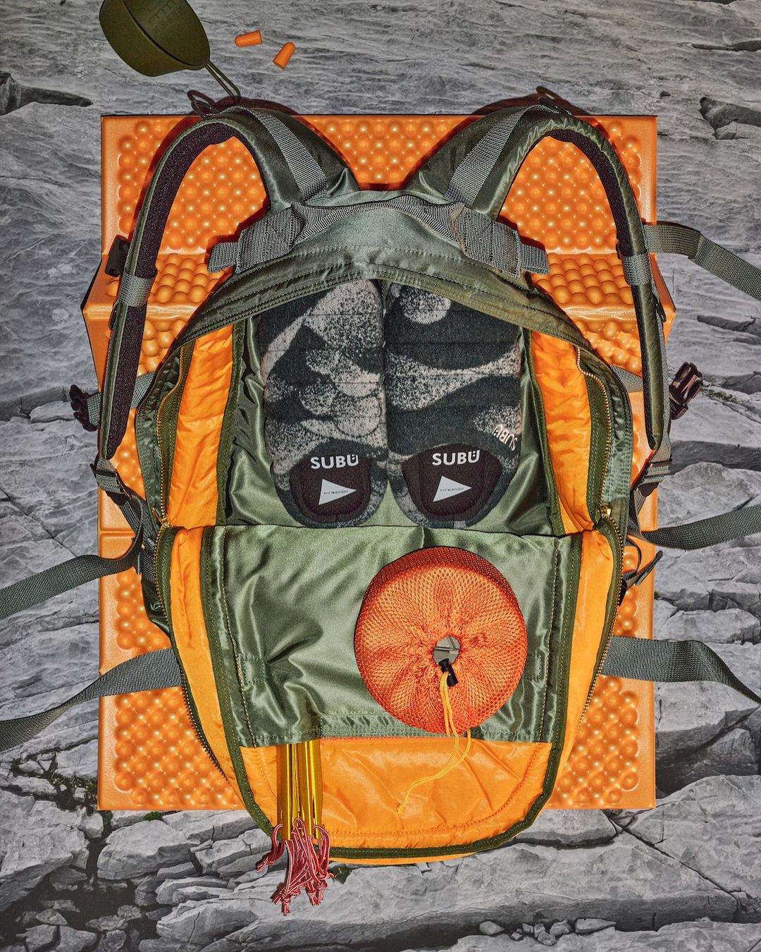 Overhead shot of an olive green backpack with orange trim, open and filled with camping gear, set on a rocky surface outdoors.