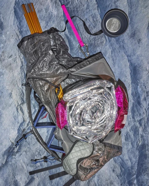 Outdoor gear laid on a textured surface: backpack, tent poles, reflective blanket, and a camping cup are neatly arranged.