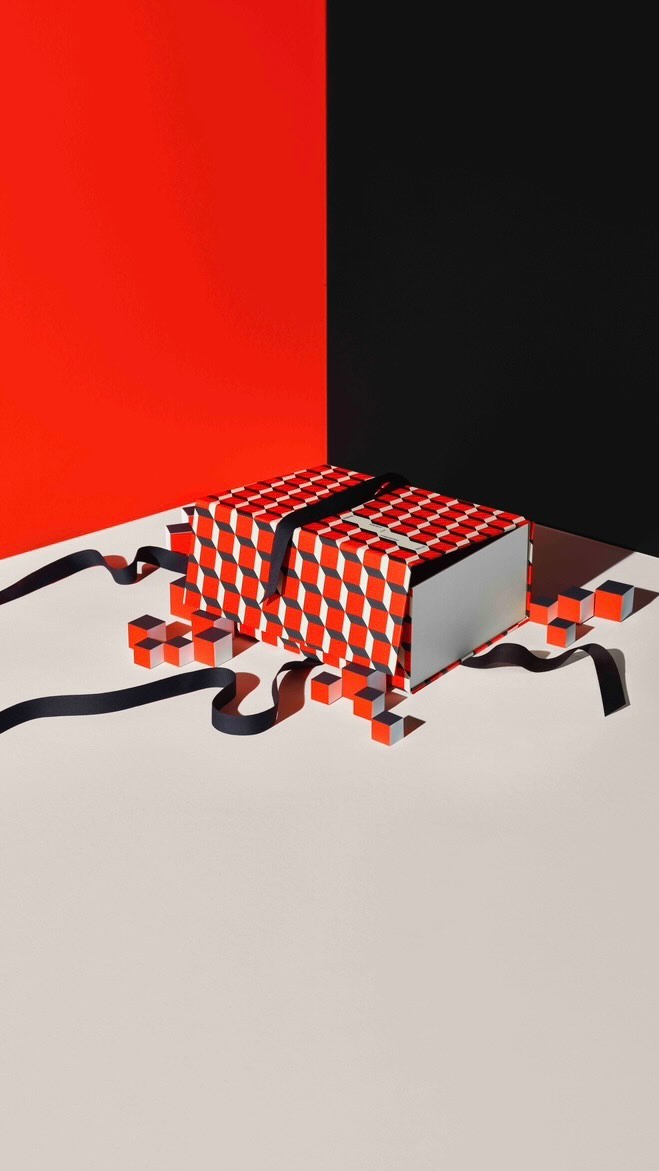 Geometric gift box with red and white pattern and black ribbon sits on a cream surface against a red and black corner backdrop.