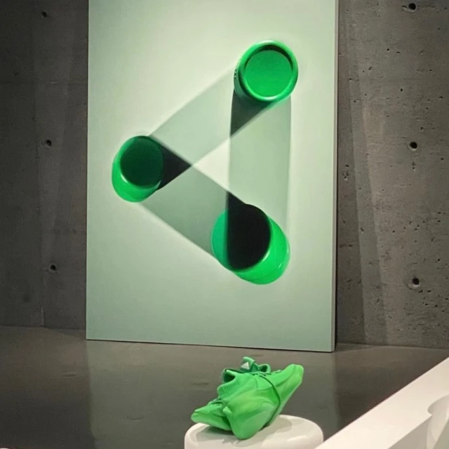 Contemporary wall art installation featuring three green cylinders casting shadows on a mint-green surface, with a green object on a pedestal below.