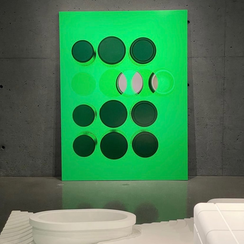 Contemporary art installation featuring a bright green geometric wall art piece with circular cutouts in an indoor gallery setting.