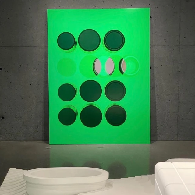 Contemporary art installation featuring a bright green geometric wall art piece with circular cutouts in an indoor gallery setting.