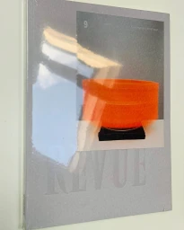 A grey magazine cover titled REVUE featuring a small inset image of an orange bowl on a dark stand with overhead reflection.