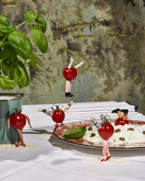 Miniature figures made from cherry tomatoes posed on a dining table with a plate of ricotta cheese and fresh basil
