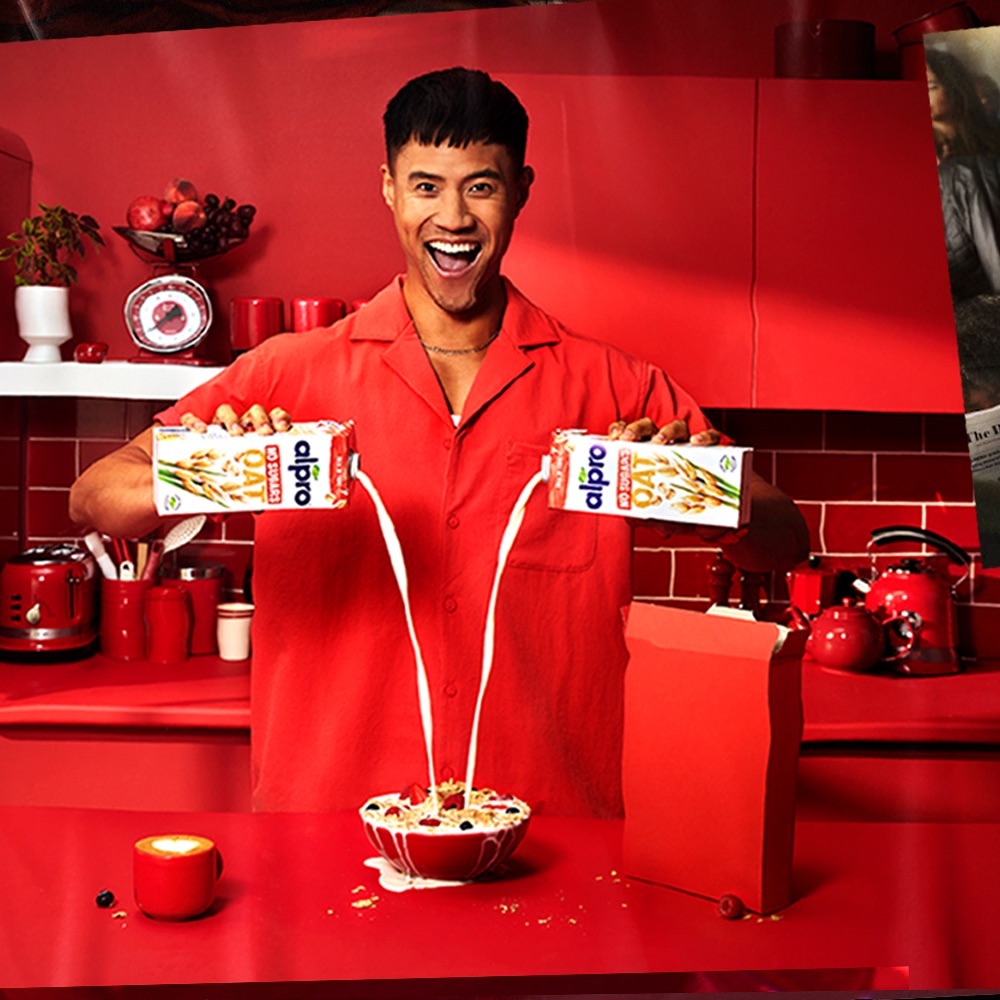 Smiling man in red kitchen pouring Alpro oat milk into bowl of cereal with berries, next to red kitchenware and Alpro carton.