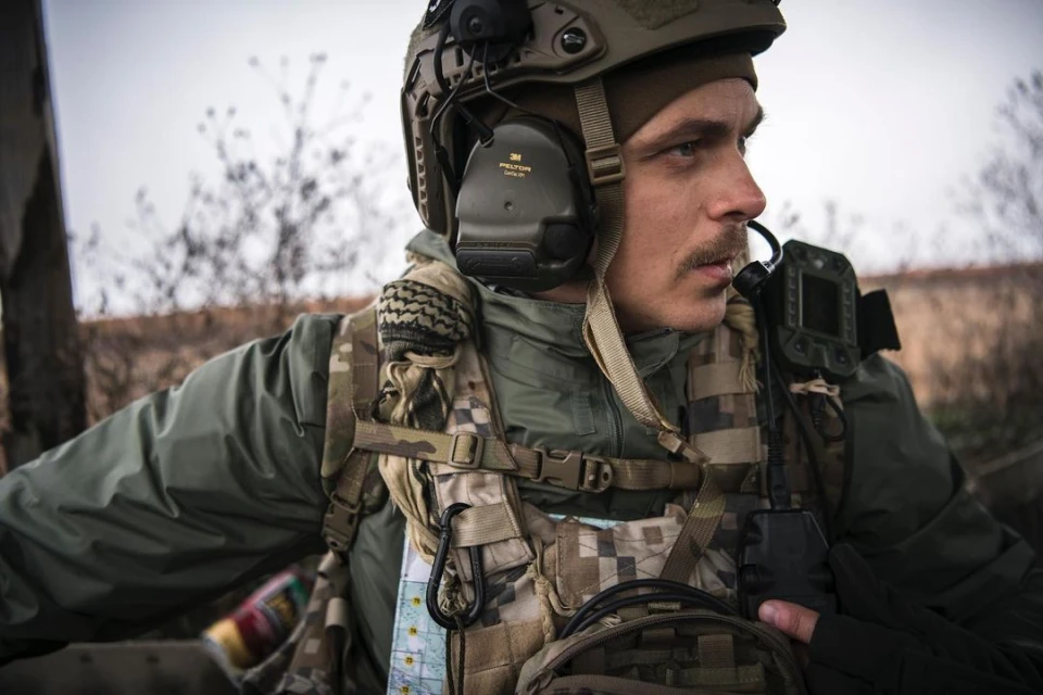 Man in military gear, helmet with headset, and tactical vest, looking off-camera in an outdoor setting with blurred vegetation.