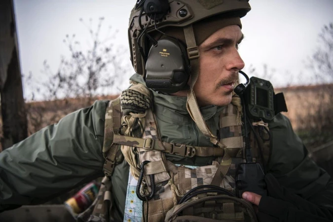 Man in military gear, helmet with headset, and tactical vest, looking off-camera in an outdoor setting with blurred vegetation.