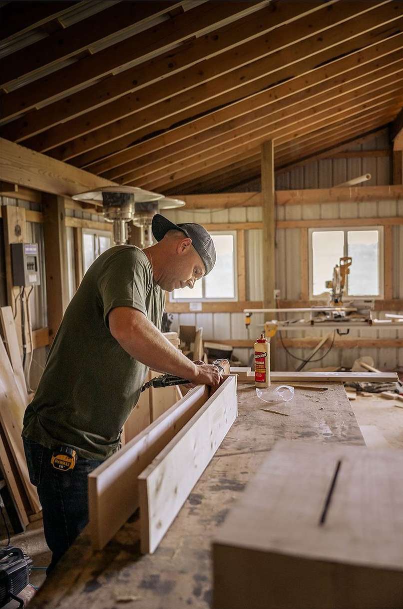 Adult man working in a woodworking shop attaching pieces of wood with a nail gun, creating a box with lumber, tools, and supplies nearby.