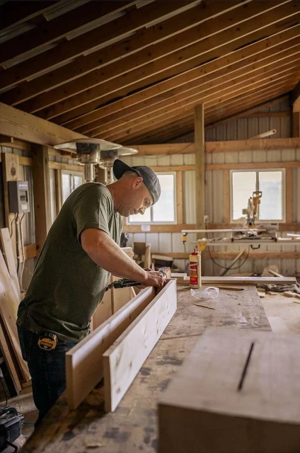 Adult man working in a woodworking shop attaching pieces of wood with a nail gun, creating a box with lumber, tools, and supplies nearby.
