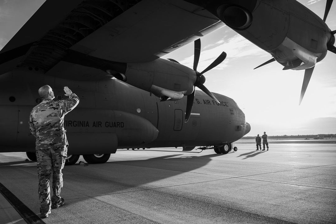 Black and white shot of a military member in uniform saluting a U.S. Air Force C-130 Hercules aircraft with two other figures nearby on a tarmac.