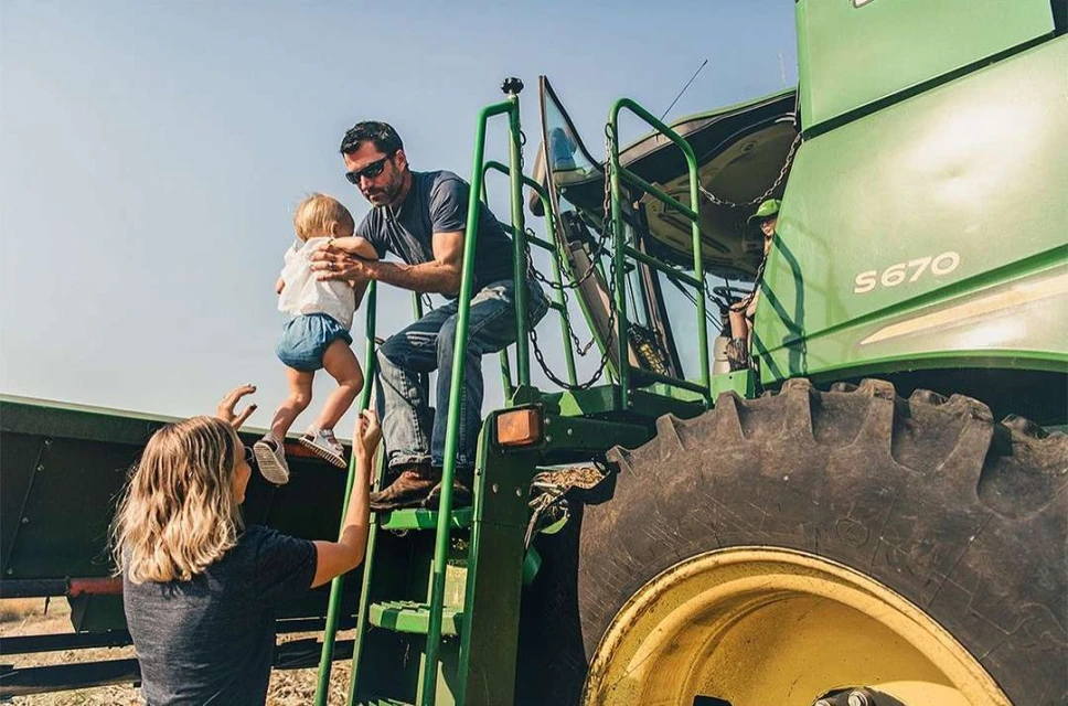 Family on a farm: a father assists a toddler descending the steps of a green John Deere S670 combine harvester, with the mother watching below.