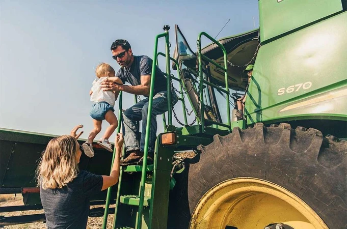 Family on a farm: a father assists a toddler descending the steps of a green John Deere S670 combine harvester, with the mother watching below.