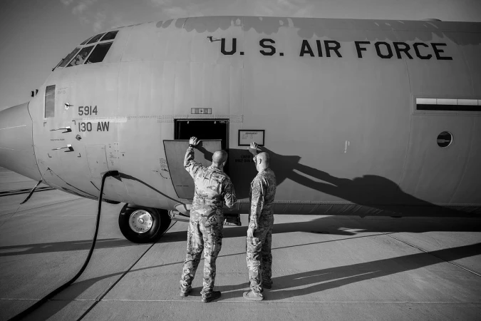 Black and white image of two military personnel in uniform standing next to a large U.S. Air Force plane on an airfield.