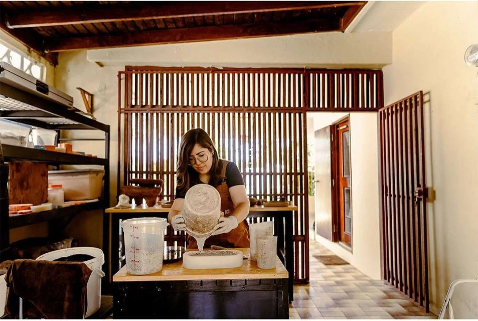 Woman with glasses and apron pouring mixture into a mold on a table in an indoor space with shelves and tiled floors.
