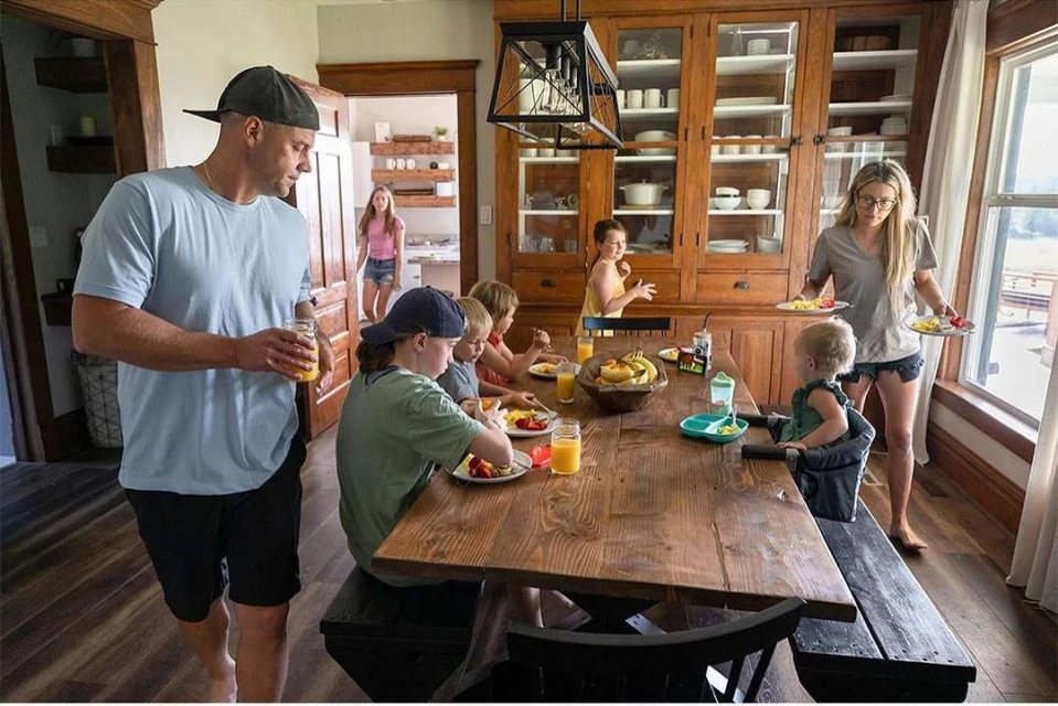 Family of seven gathers around a wooden dining table for breakfast, with fruit, juice, and dishes visible in an indoor setting.