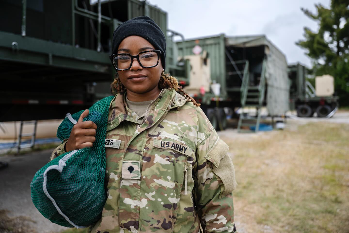 Portrait of an adult female in military fatigues, wearing glasses and a beanie, holding a green mesh bag against a blurred military backdrop.