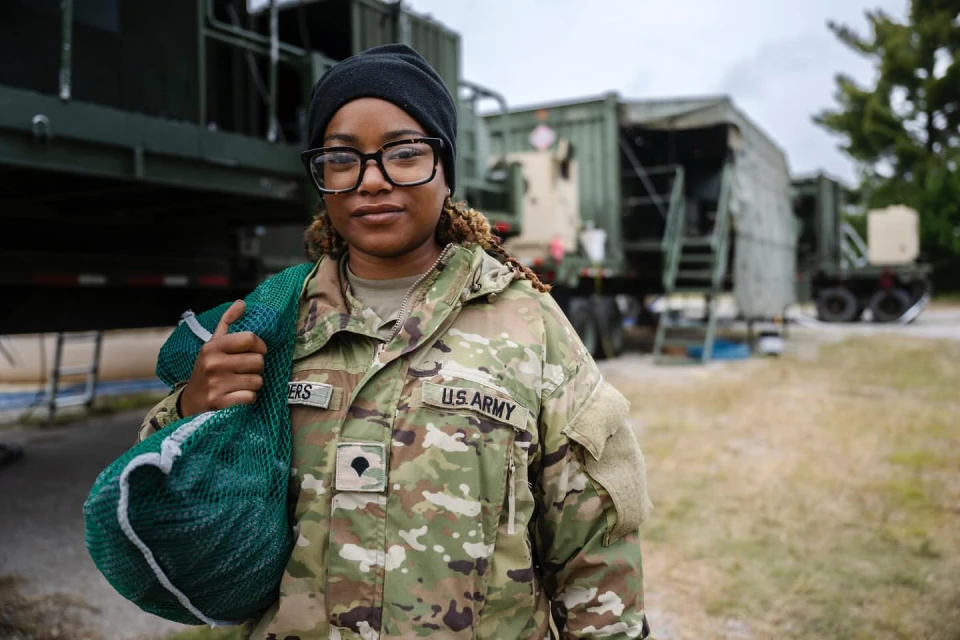 Portrait of an adult female in military fatigues, wearing glasses and a beanie, holding a green mesh bag against a blurred military backdrop.