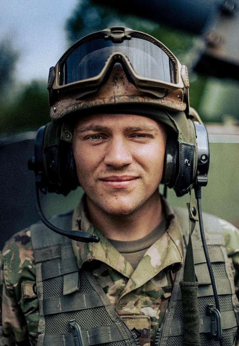 Close-up portrait of a military member in camouflage helmet, goggles, vest, and headphones smiling at the camera outdoors.
