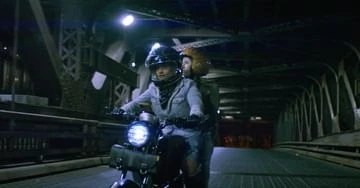 Two people riding a motorcycle at night under a metal bridge, illuminated by the headlight in a medium shot with dark mood.