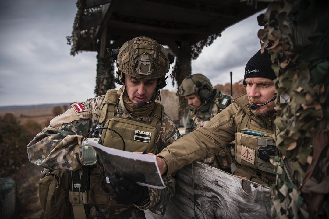 Three soldiers in camouflage gear consult a map outdoors, two wearing helmets and one a beanie, in a military or training context.