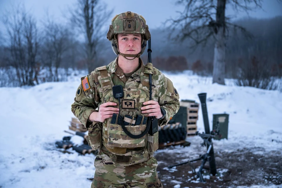 Full shot of a young adult male soldier in camouflage uniform and helmet posing in a snowy, wooded outdoor environment with equipment.