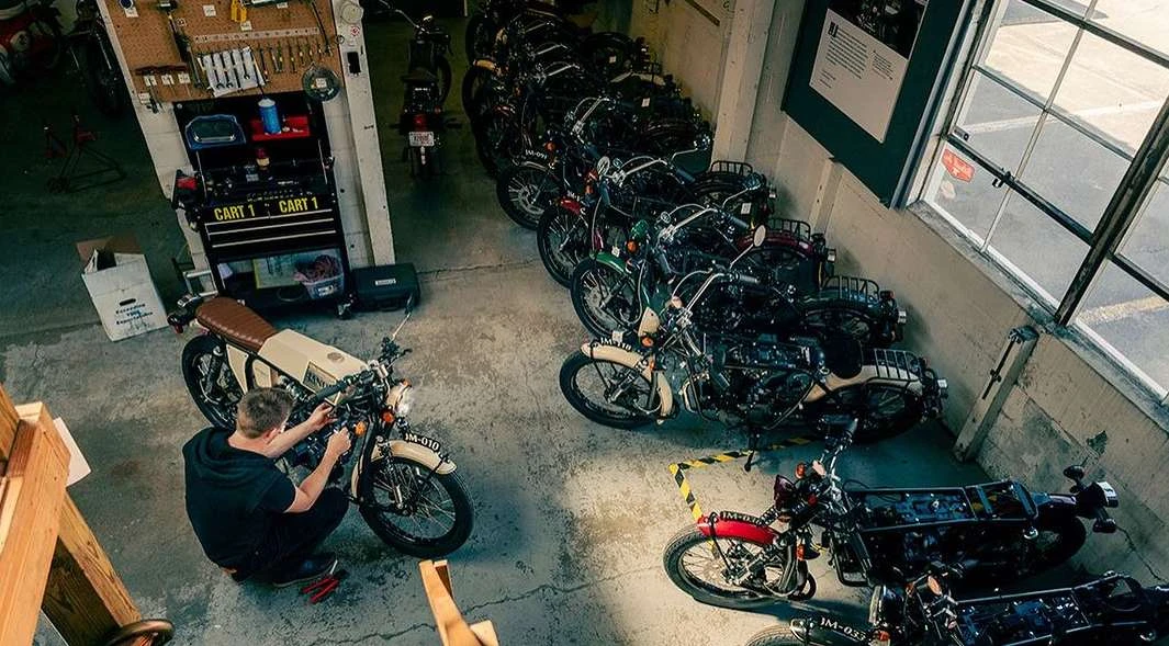 Mechanic repairs a vintage motorcycle in a repair shop surrounded by a collection of motorcycles; shot from a high angle.