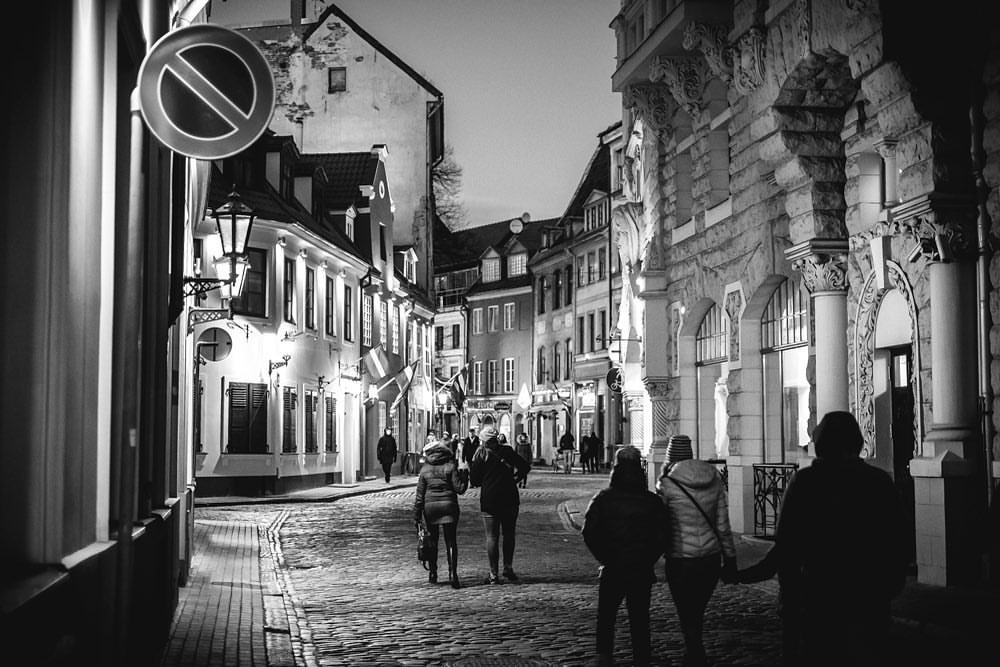 Monochrome image of people walking along cobblestone street flanked by old buildings in European city at night with street lights.
