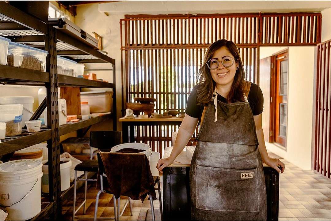 Smiling woman wearing an apron standing in a sunlit pottery studio, posing next to a table with storage shelving and pottery in the background.