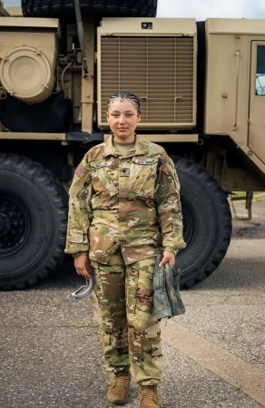 Full shot of a woman in camouflage military uniform standing in front of a large military vehicle with large tires and cloudy skies.