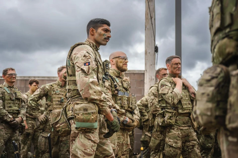 A group of soldiers in camouflage uniforms stand outdoors under a cloudy sky. They are in formation, looking towards the left side of the frame.