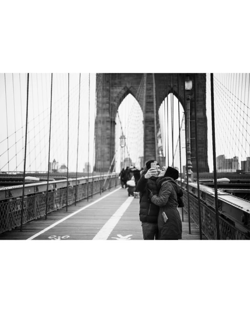 Black and white photo of a couple taking a selfie on the Brooklyn Bridge, with the bridge's architecture and cityscape in the background.