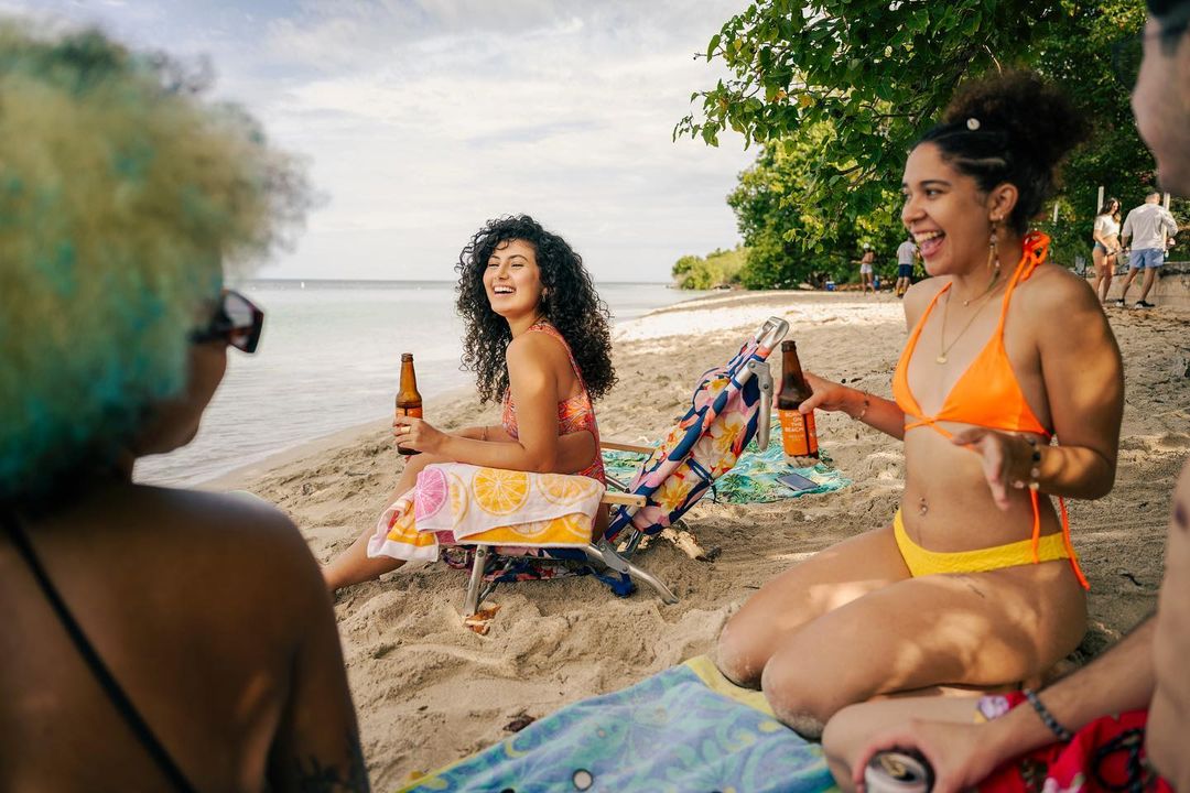 Four people relaxing on a sandy beach, two women with curly hair hold beer bottles, wearing swimsuits, next to trees and ocean views.