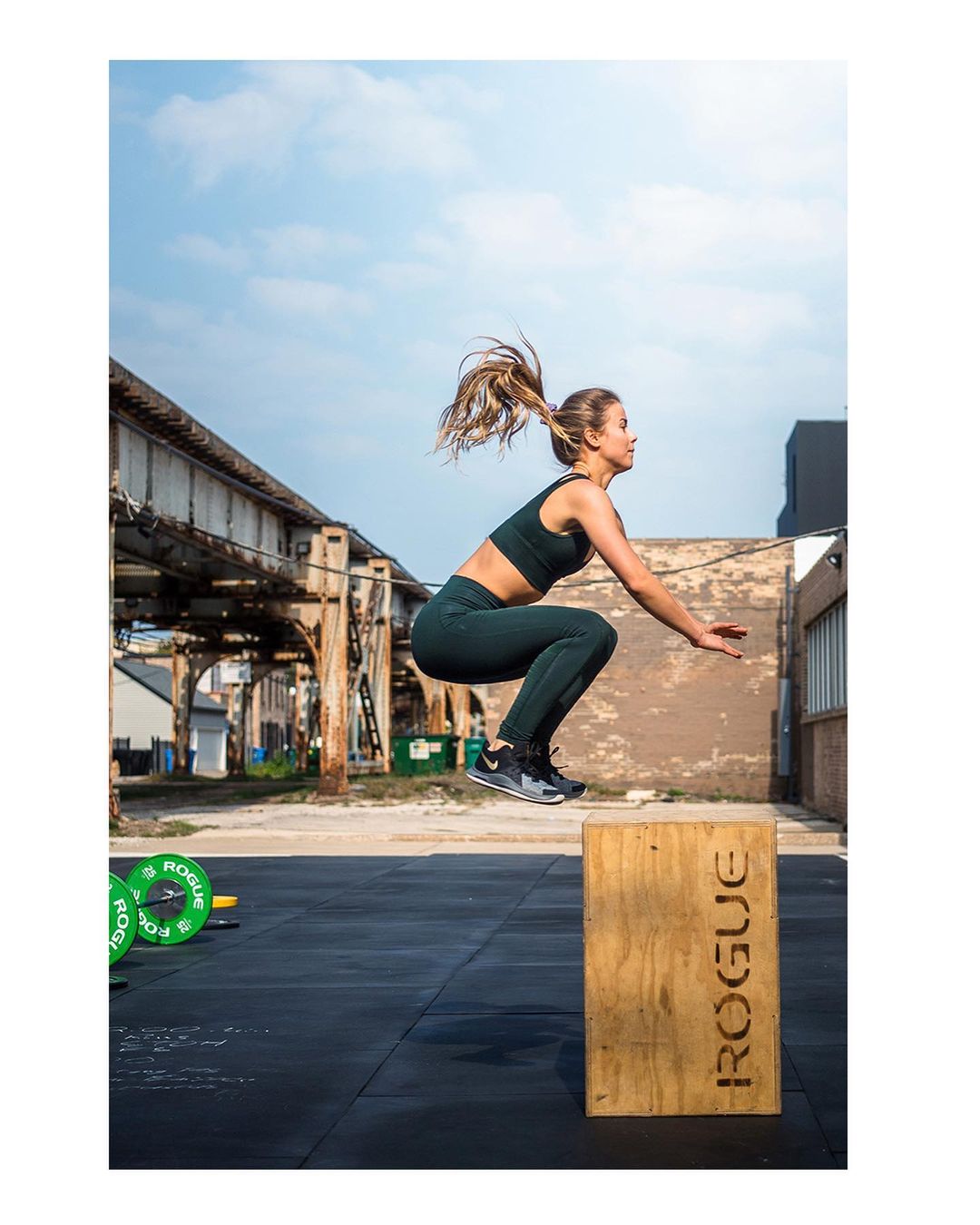 Athletic woman in sports bra and leggings jumps onto wooden box during outdoor workout against urban backdrop with green weights.