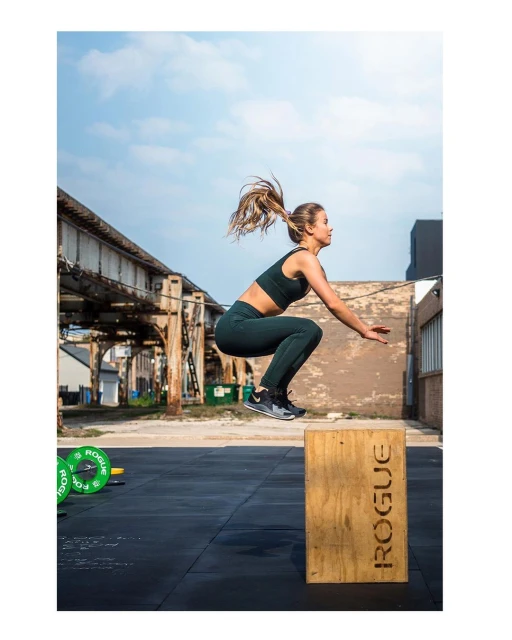 Athletic woman in sports bra and leggings jumps onto wooden box during outdoor workout against urban backdrop with green weights.
